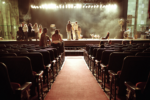 An auditorium with a red carpet aisle leading to a stage. Several people are gathered on the stage under bright stage lights, while others sit in the audience seats.