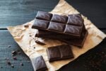 Dark chocolate bars with cocoa leaf designs on top, stacked on brown paper with chocolate shavings scattered around.